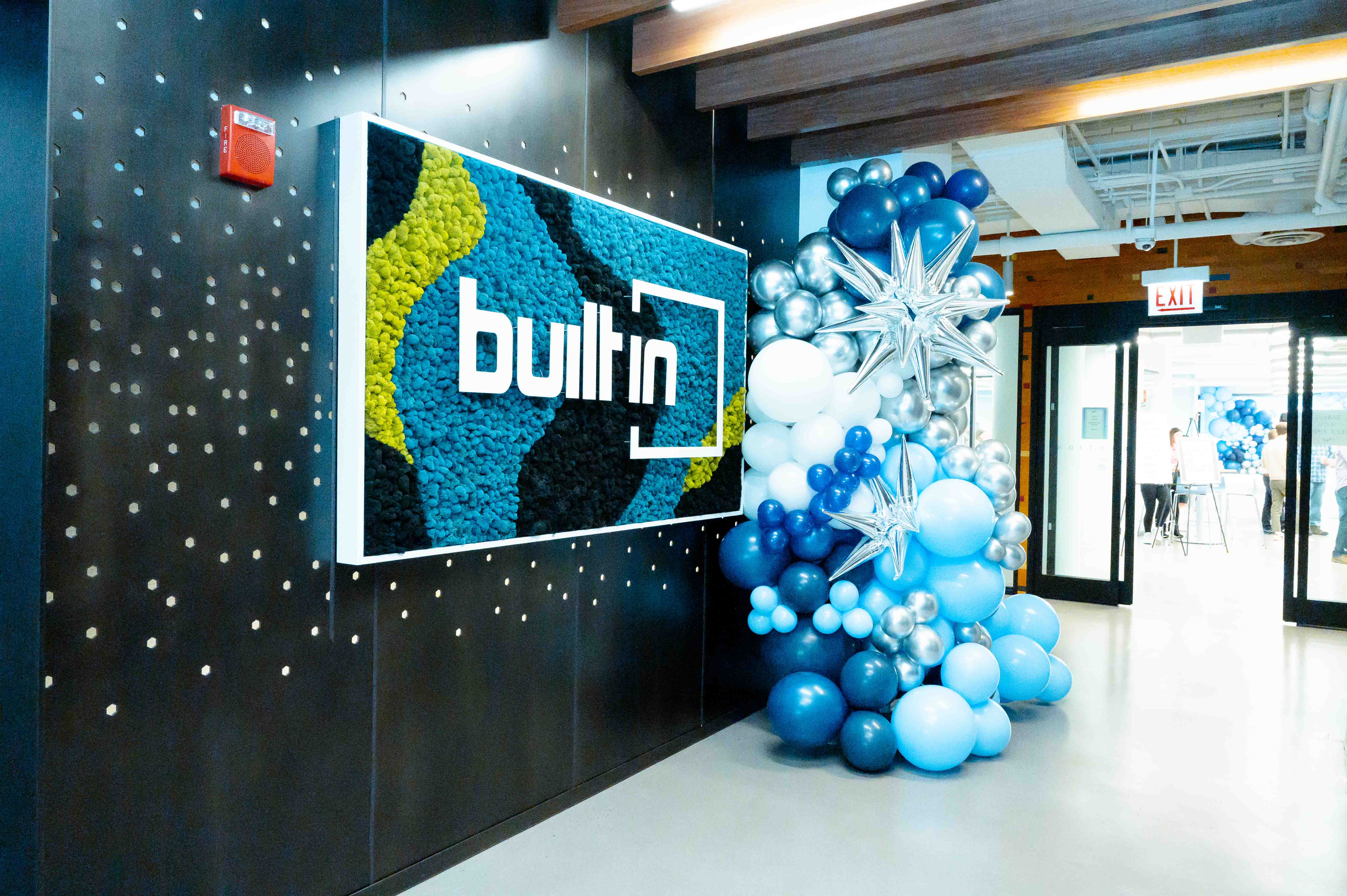 Artwork featuring the Built In logo against multicolored moss on the wall next to a bouquet of white, silver and blue balloons.