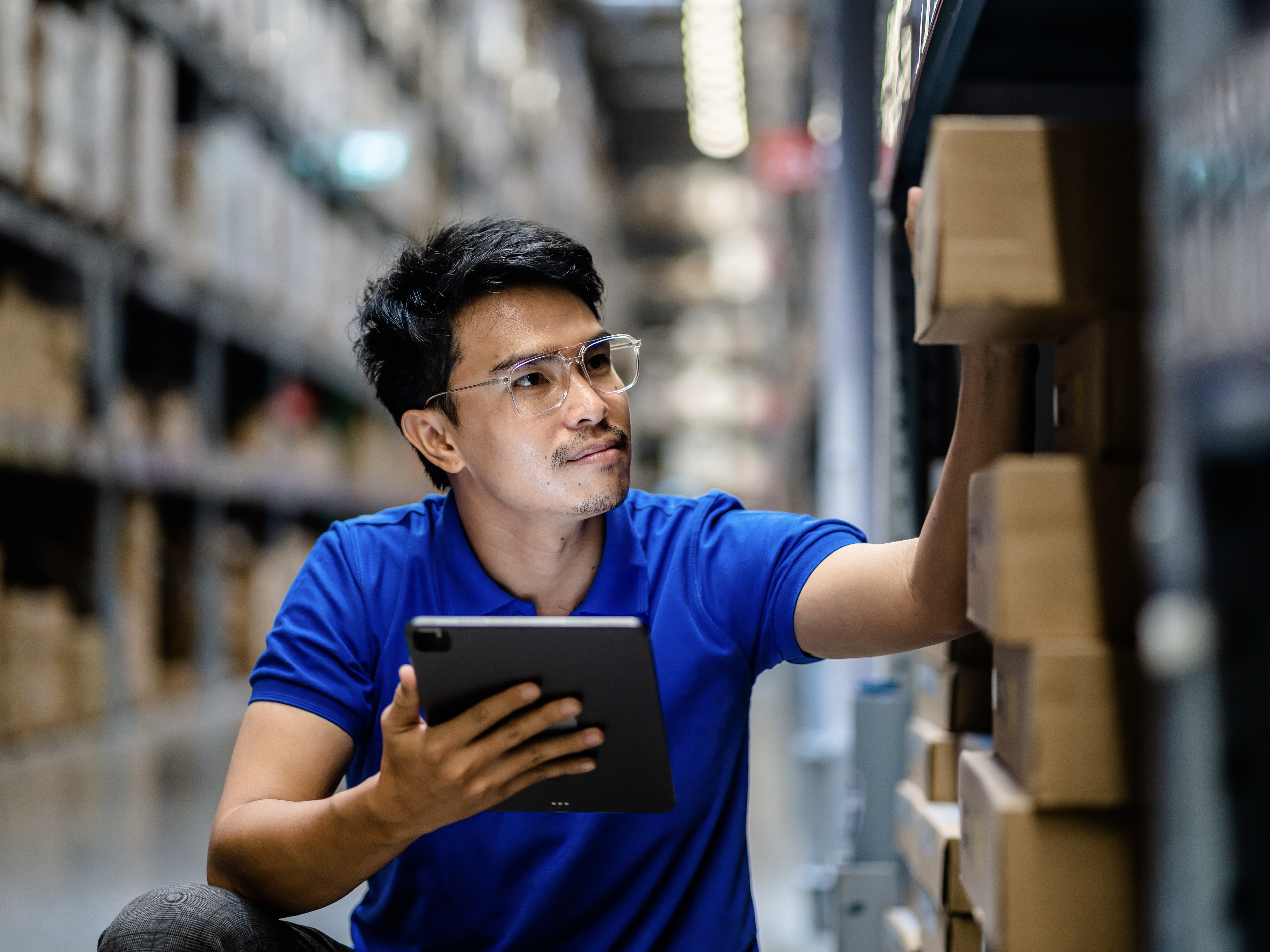 A warehouse worker consults a tablet as he inspects boxes on a shelf.