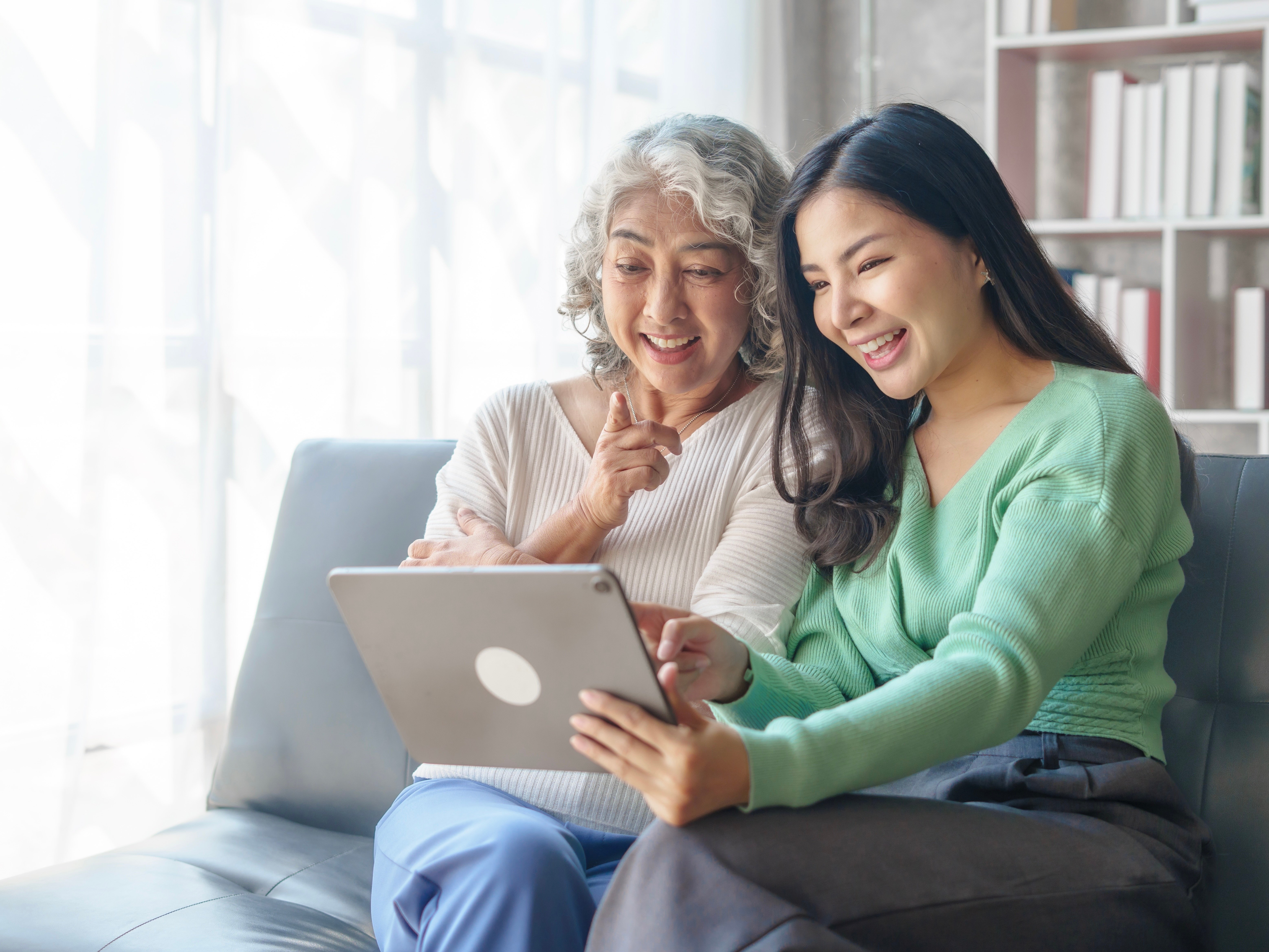 An older and younger woman smile and gesture at a tablet in front of them.