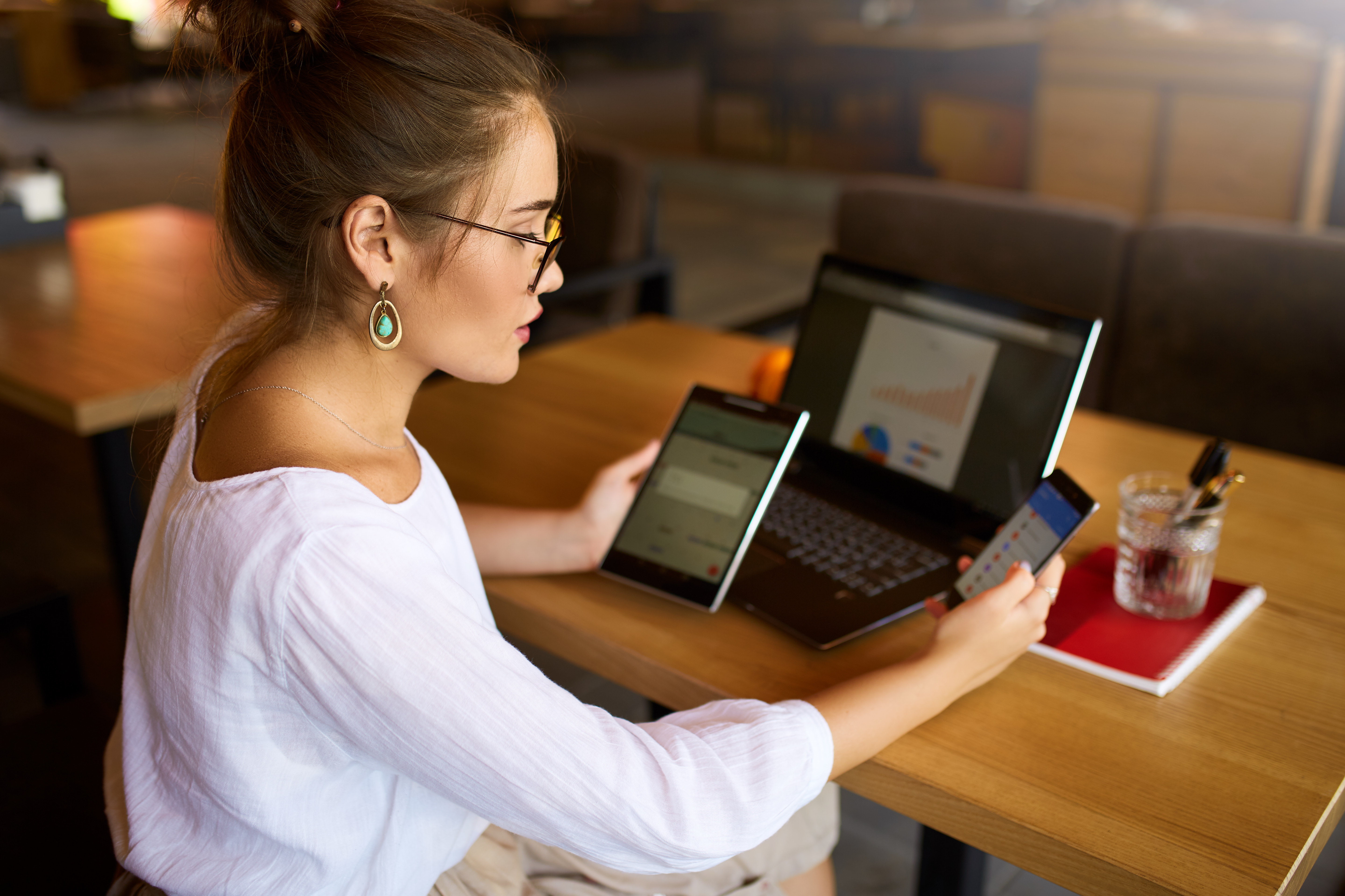 A woman looks between her cell phone, tablet, and laptop as she works at a table.