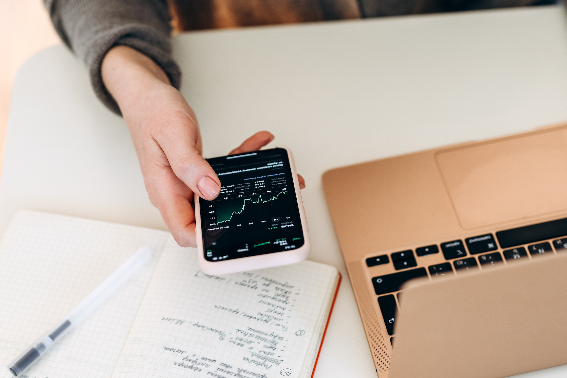 Person's hand holding a smart phone and looking at investment performance. They sit at a white desk with a gold macbook and a notebook. 